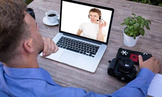 Man in a blue shirt video-chats on a laptop at an outdoor table, with a camera, coffee, plant, and notes nearby.