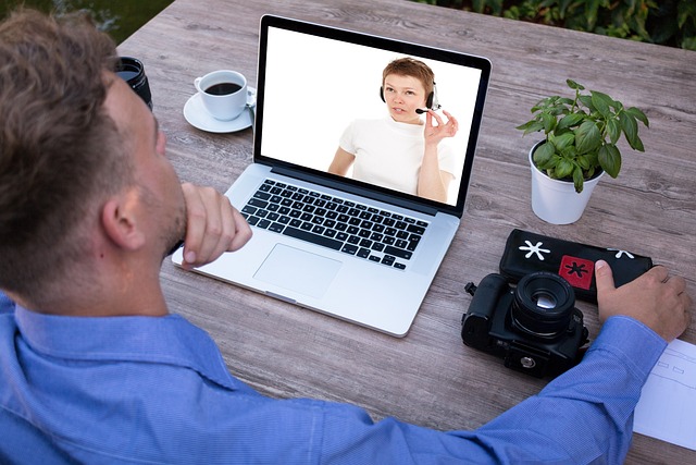 Man in a blue shirt video-chats on a laptop at an outdoor table, with a camera, coffee, plant, and notes nearby.