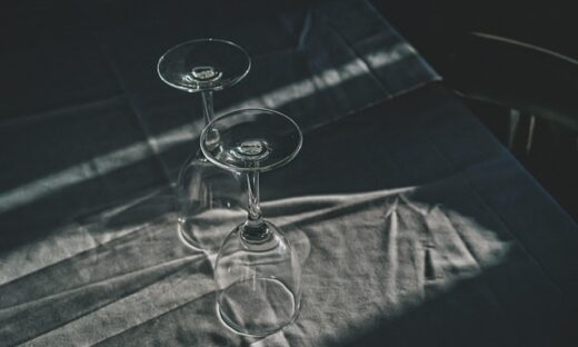 Two empty wine glasses balanced on a dark table with dramatic light and shadows on the fabric surface. Clean, moody still life.