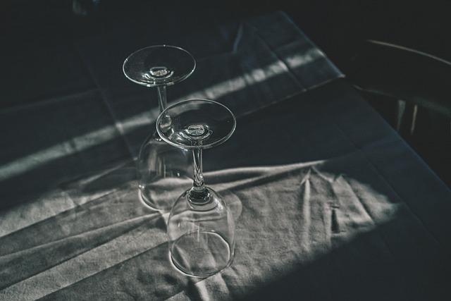 Two empty wine glasses balanced on a dark table with dramatic light and shadows on the fabric surface. Clean, moody still life.