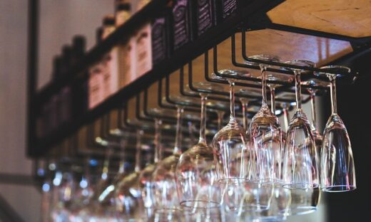 Row of wine glasses hanging upside down from a bar rack with bottles on a shelf above (background blurred).