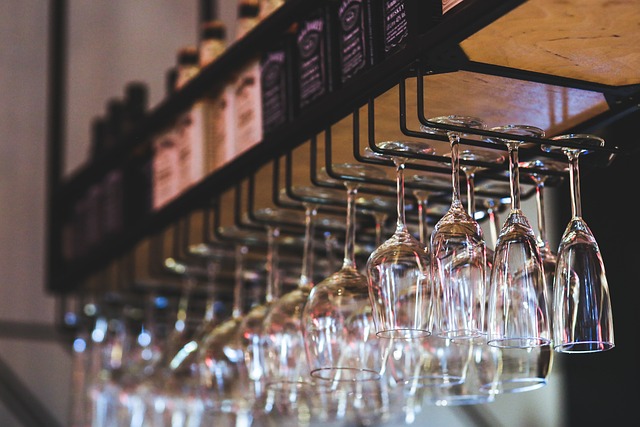 Row of wine glasses hanging upside down from a bar rack with bottles on a shelf above (background blurred).