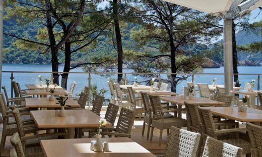 Outdoor lakeside cafe with beige tables and woven chairs, overlooking a blue lake and forested hills.