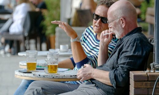 Two men sit at an outdoor cafe table with beer, one wearing sunglasses and striped shirt, gesturing as they chat.