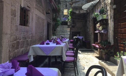Outdoor street-side restaurant in a narrow stone alley, with tables covered in purple linens and purple chairs, and potted plants along the walls.
