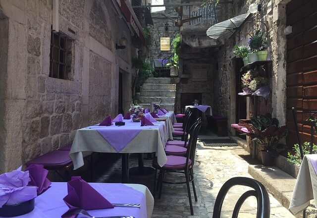 Outdoor street-side restaurant in a narrow stone alley, with tables covered in purple linens and purple chairs, and potted plants along the walls.
