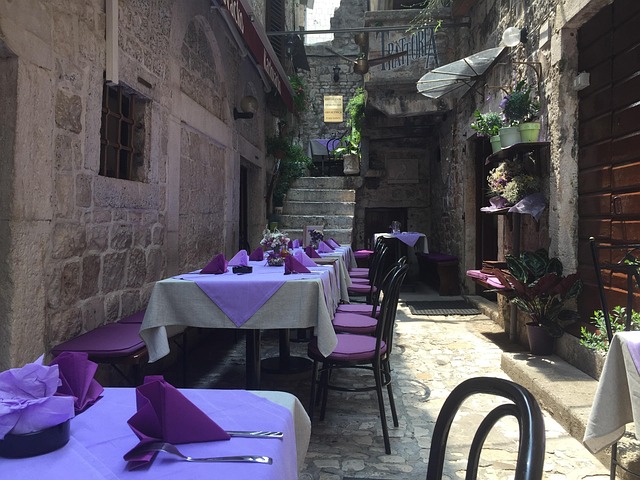 Outdoor street-side restaurant in a narrow stone alley, with tables covered in purple linens and purple chairs, and potted plants along the walls.