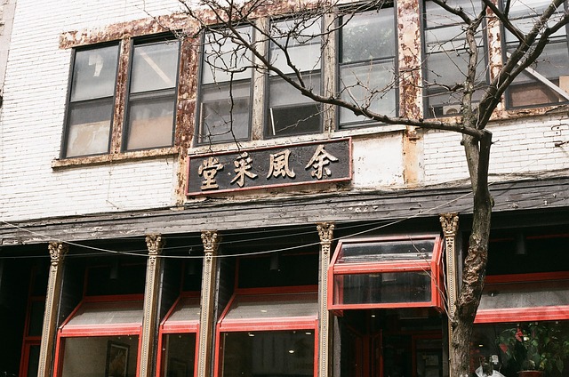Old storefront with peeling paint, rusted sign, and red-framed windows, on a tree-lined street.