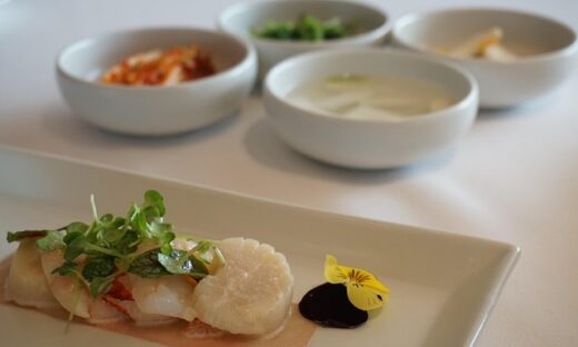 Sliced white fish with microgreens on pale sauce, garnished with a yellow edible flower and a dark sauce on a rectangular plate.