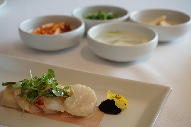 Sliced white fish with microgreens on pale sauce, garnished with a yellow edible flower and a dark sauce on a rectangular plate.